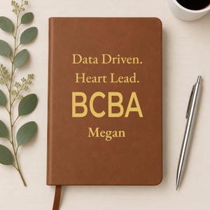 May include: A brown notebook with a gold-colored inscription that reads Data Driven. Heart Lead. BCBA Megan. The notebook features a smooth, solid-colored cover and a matching brown ribbon bookmark extending from the bottom edge of the front cover.