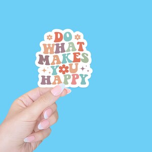 May include: A white sticker with the text "DO WHAT MAKES YOU HAPPY" in colorful, retro-style lettering. The sticker is being held by a hand against a light blue background.