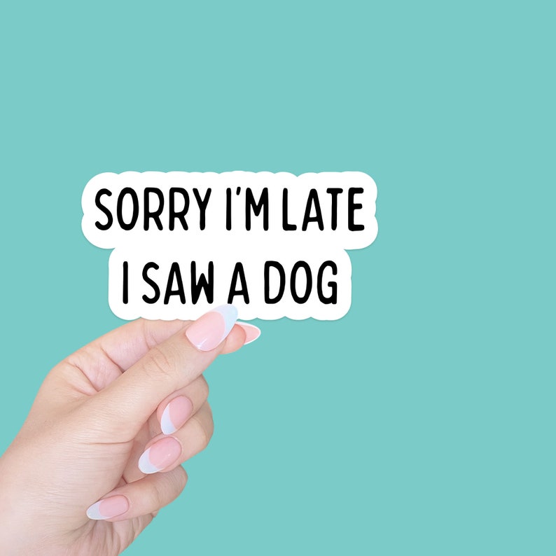 May include: A white sticker with black text that reads "Sorry I'm late I saw a dog". A hand is holding the sticker in front of a turquoise background.