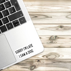 May include: A silver laptop computer with a white sticker on the top right corner that says "Sorry I'm late I saw a dog". The laptop is on a light brown wooden surface.
