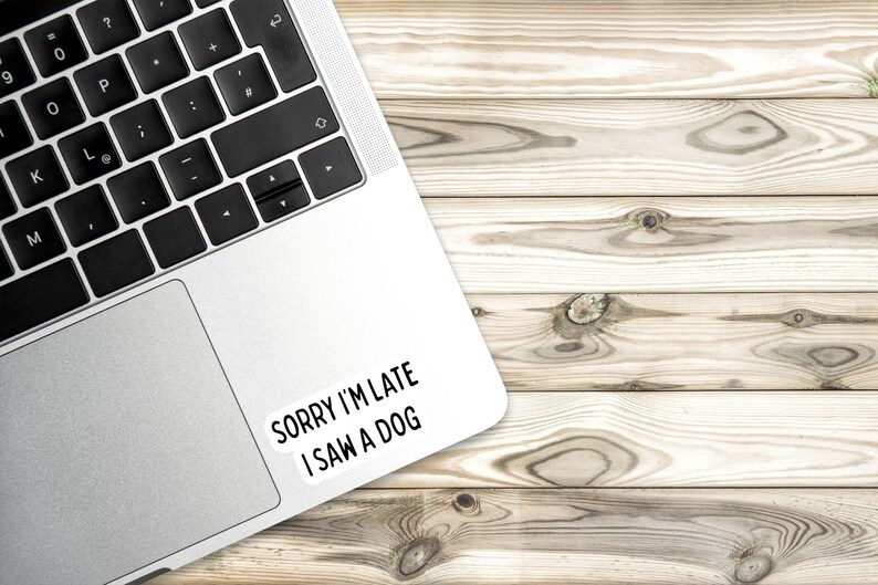 May include: A silver laptop computer with a white sticker on the top right corner that says "Sorry I'm late I saw a dog". The laptop is on a light brown wooden surface.