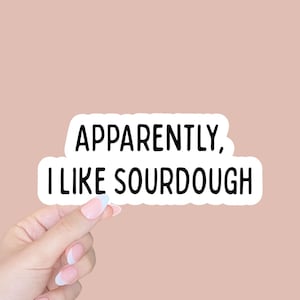 May include: A white sticker with the text "APPARENTLY, I LIKE SOURDOUGH" in black, held by a hand with manicured nails against a pink background. The sticker has a rounded rectangular shape.