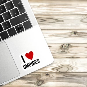 Może przedstawiać: Srebrny laptop z białą naklejką z napisem "Kocham sędzi&oacute;w" z czerwonym sercem. Laptop leży na jasnobrązowej drewnianej powierzchni.
