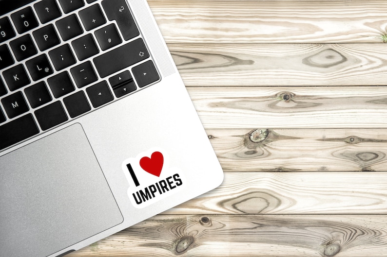Może przedstawiać: Srebrny laptop z białą naklejką z napisem "Kocham sędzi&oacute;w" z czerwonym sercem. Laptop leży na jasnobrązowej drewnianej powierzchni.