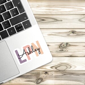 May include: A sticker with the letters LPN in pink, orange, and purple with the name Ashley written in cursive underneath.
