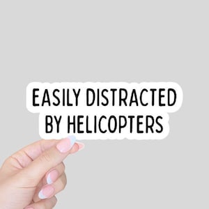 May include: A white sticker with the text "EASILY DISTRACTED BY HELICOPTERS" in black, held by a hand with manicured nails. The sticker has a rounded rectangle shape.