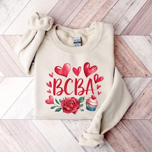 May include: A beige sweatshirt with a red heart design and the text "BCBA" in red. The design also includes a red rose and a cupcake with red frosting.