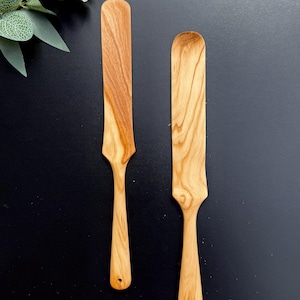 Puede incluir: Dos espátulas de madera de color marrón claro con vetas visibles. Cada utensilio presenta una cabeza larga, plana y rectangular que se estrecha hacia un mango delgado con un pequeño orificio circular en el extremo inferior para colgar.