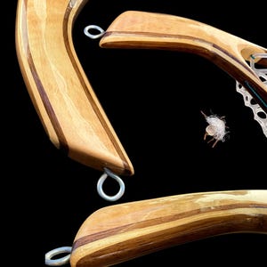 Puede incluir: Dos mangos de madera curvada para redes de pesca con laminados de grano de madera claro y oscuro, acabado pulido y ojales de metal plateado en los extremos. El texto "Heirloom, Handcrafted Net • Handle Details" aparece impreso en la parte inferior.