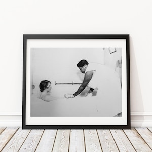 May include: A black and white photograph of two people in a bathroom. One person is sitting in a bathtub and the other person is standing and washing the person in the tub.