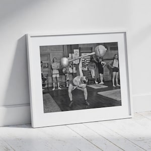 May include: Black and white framed print of a weightlifter in a gym, lifting a barbell overhead. Other people are in the background, observing. The image is in a white frame, leaning against a white wall and a wooden floor.