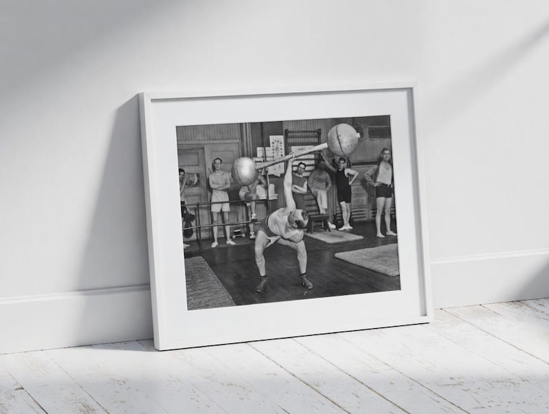 May include: Black and white framed print of a weightlifter in a gym, lifting a barbell overhead. Other people are in the background, observing. The image is in a white frame, leaning against a white wall and a wooden floor.