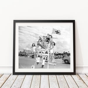 Könnte beinhalten: Ein Schwarzweißfoto einer Gruppe von Menschen, die auf einem Rettungsschwimmerstand an einem Strand stehen. Die Menschen tragen alle Badeanzüge und lächeln. Im Hintergrund weht eine Rotkreuzflagge.