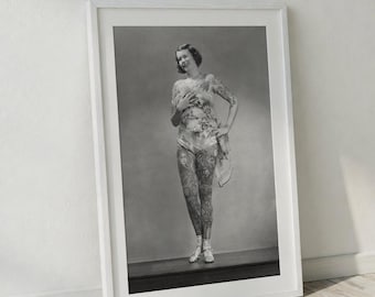 Puede incluir: Impresión enmarcada en blanco y negro de una mujer con extensos tatuajes en brazos y piernas. Viste ropa clara y está de pie sobre una superficie oscura. La imagen está apoyada contra una pared blanca.