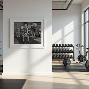 May include: A framed black and white print of a weightlifting competition hangs on a white wall in a gym. The gym features weight racks, dumbbells, and a stationary bike. Natural light streams in from large windows, illuminating the space.