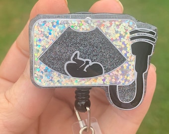 Peut inclure: Un porte-badge noir et argenté à paillettes avec une image d'échographie en noir et blanc d'un bébé à l'intérieur. Le porte-badge a un cordon noir et blanc et un clip noir.