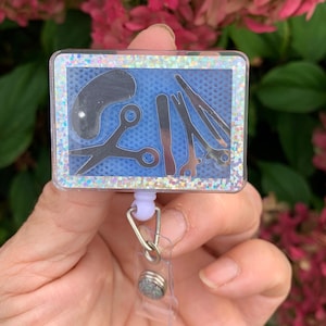 May include: A blue and silver glitter badge holder with a clear plastic insert featuring a silhouette of hairdressing tools. The holder is attached to a clear plastic retractable badge reel.