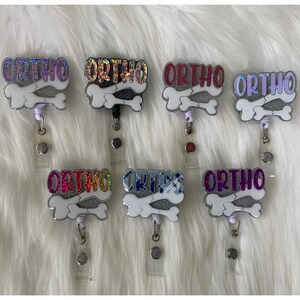 May include: Six different coloured badge reels with the word "ORTHO" written on them. The badge reels are decorated with glitter and have a bone design.
