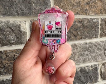 Puede incluir: Encanto de bolsa IV de resina rosa con purpurina con una botella de poción de amor, corazones y el texto "LOVE POTION" y "XOXO".