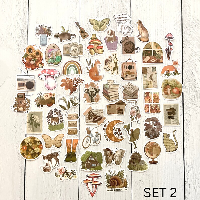 Op de afbeelding: Een set van 40 kleurrijke stickers met verschillende illustraties, waaronder paddenstoelen, vlinders, een slak, een huis, een fiets, een hert, een vos, een kat, een kikker, een camera, een cassettedeck, een regenboog, een maan, een globe, een koffer, een boek, een raam, een ladekast en een paar laarzen. De stickers zijn op een witte houten achtergrond. De tekst "SET 2" staat in de rechterbenedenhoek van de afbeelding.