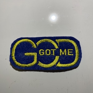 May include: A blue embroidered patch with the words "GOD GOT ME" in yellow thread. The patch has a rectangular shape with rounded corners. The text is centered on the patch.