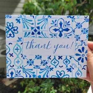 May include: A white card with a blue tile pattern and the words "thank you!" in cursive script. The card features a repeating design of blue floral and geometric shapes, with a watercolor effect. The card is held in a hand.