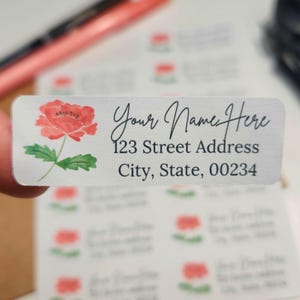 Op de afbeelding: Rechthoekige adreslabels met een aquarel klaproosontwerp. De labels bevatten de tekst "Your Name Here", adresgegevens en een postcode. De klaproos is rood met groene bladeren.