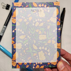 May include: A floral patterned notepad with the word "NOTES" at the top. The notepad has a blue border with orange flowers and green leaves. The background of the notepad is white with a floral pattern. A black pen, blue pen, and binder clip are visible.