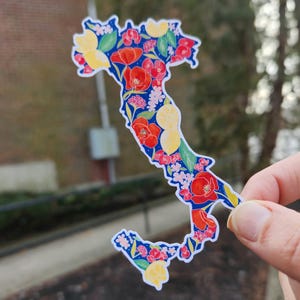 May include: A sticker in the shape of Italy, adorned with a floral design. The pattern includes red poppies, yellow lemons, and pink flowers set against a dark blue backdrop. The sticker is held by a hand.