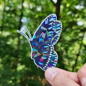 May include: A holographic butterfly sticker with a geometric design. The butterfly's wings are filled with blue, purple, green, and pink shapes. The sticker is held against a blurred green background.