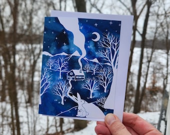 以下が含まれることがあります： 夜に雪が降った森の中に座っている白いウサギの水彩画です。ウサギは三日月と空の星を見上げています。背景には小さな家が見えます。