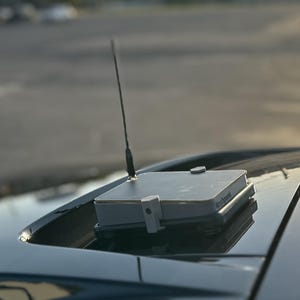 May include: A square, light grey electronic device with a thin, vertical black antenna mounted on the roof of a black vehicle. The device features a small circular knob on its top surface and is positioned within the open sunroof area of the car.