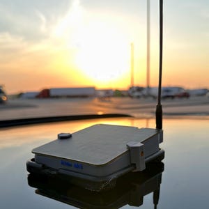 May include: A rectangular electronic device with a grey textured top surface and a vertical black antenna. The side of the device features the blue text "Kilo Mk8" and is secured by light grey plastic clips on a reflective surface.