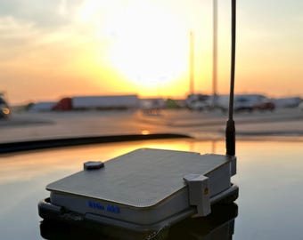 May include: A rectangular electronic device with a grey textured top surface and a vertical black antenna. The side of the device features the blue text "Kilo Mk8" and is secured by light grey plastic clips on a reflective surface.