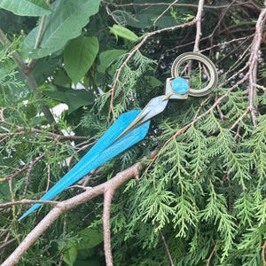 Peut inclure: Une paire de ciseaux décoratifs avec une lame bleu turquoise et une poignée dorée. Les ciseaux comportent une pierre précieuse bleu clair et sont placés sur un fond de feuillage vert.
