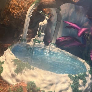 May include: A miniature decorative fountain with a statue of a deer, surrounded by arches and faux greenery. The fountain has a blue water feature and is set in a terrarium environment. The overall design is reminiscent of a classical garden.