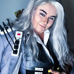 以下が含まれることがあります： ライトブルーのブレザー、黒いベスト、白い襟付きシャツを着たコスプレ衣装を着た人。衣装には肩に取り付けられた電源タップとベストの黄色のブロックが含まれています。