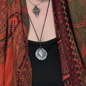 Peut inclure: Un collier pendentif argenté avec un motif de visage détaillé, suspendu à un cordon noir. Un deuxième collier argenté avec un pendentif décoratif est également visible. Les colliers sont portés sur un haut noir et un châle orange et rouge à motifs.