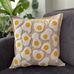 May include: A beige throw pillow with a white and yellow embroidered design of multiple fried eggs.