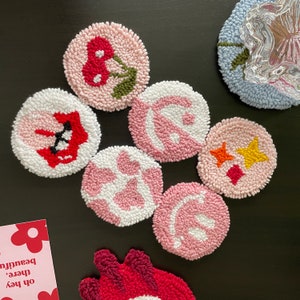 May include: Seven pink and white round crocheted coasters with various designs. The designs include a cherry, a swirl, a star, a smiley face, and a geometric pattern.