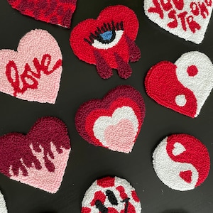 May include: A collection of 11 handmade felt heart-shaped and round ornaments with various designs. The designs include a red heart with white polka dots, a red heart with a white heart inside, a red heart with a white yin-yang symbol, a red heart with a white eye, a red heart with white dripping paint, a red heart with the word "love" in white, a white heart with the words "you're strong" in red, a white heart with a red yin-yang symbol, a white heart with a red smiley face, a white heart with a red eye, and a red heart with a white dripping paint.