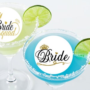 May include: Two margarita glasses with white paper circles on them. The circle on the left glass says "Bride Squad" in black with a gold crown and ring. The circle on the right glass says "Bride" in black with a gold crown and heart. Both glasses have a lime wedge on the rim.