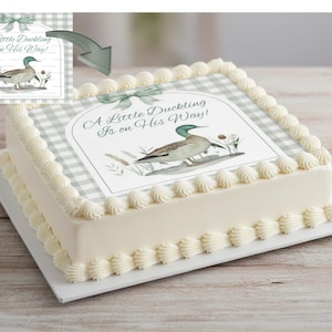 May include: A rectangular cake with white frosting and decorative piped edges. The top features a light green and white checkered pattern with a printed image of a duck and the text "A Little Duckling Is on His Way!" A light green bow is at the top.