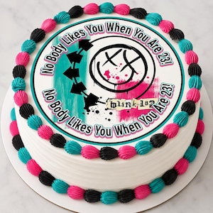 May include: A round cake with white frosting, decorated with alternating pink, black, and teal piped borders. The top features a circular graphic with the text "No Body Likes You When You Are 23!" and "blink-182" alongside a black smiley face logo.