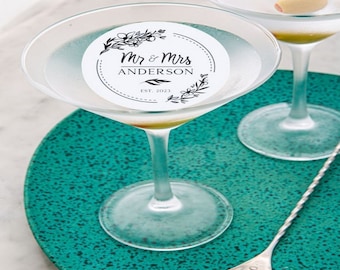 May include: Two martini glasses with a white sticker on each. The sticker has a black floral design and the text "Mr. & Mrs. ANDERSON EST. 2023". The glasses are on a teal plate with a silver cocktail stirrer.