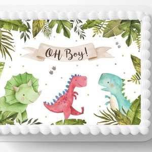 May include: A rectangular cake decorated with a watercolor design featuring three friendly dinosaurs in green, red, and blue. The phrase "OH BOY!" is written on a banner above the dinosaurs, surrounded by green foliage and a white frosting border.