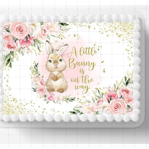 May include: A white cake with a pink and gold frosting design. The cake is decorated with a watercolor illustration of a bunny with the text "A little bunny is on the way".