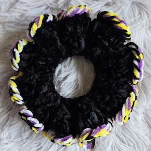 May include: A black scrunchie with a non-binary pride flag color scheme. The scrunchie is made of a soft, plush material and features yellow, white, purple, and black accents. The scrunchie is circular and has a hole in the center.