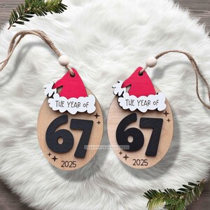 May include: Two wooden Christmas ornaments with a Santa hat design. The ornaments are oval-shaped with the text "THE YEAR OF" above the number "67" and the year "2025". Each ornament has a jute string for hanging.
