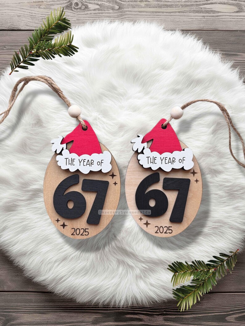 May include: Two wooden Christmas ornaments with a Santa hat design. The ornaments are oval-shaped with the text "THE YEAR OF" above the number "67" and the year "2025". Each ornament has a jute string for hanging.